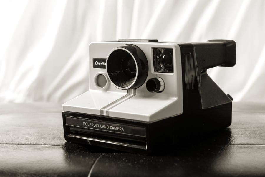 A boxy Polaroid-style instant camera with a large square print ejecting from the front